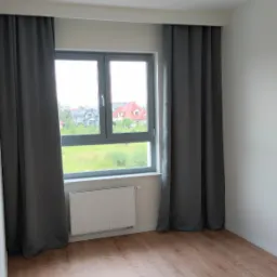 Pusty pokój z nową podłogą z paneli, szare zasłony przy oknie, białe ściany i grzejnik pod oknem, oświetlenie sufitowe na szynie.