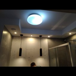 VPBud - Łazienka w trakcie remontu z zaznaczonymi wymiarami na ścianie, widoczne lampy wiszące i kabina prysznicowa.