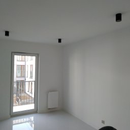 VPBud - Pusty, pomalowany na biało pokój z błyszczącą podłogą, czarnymi reflektorami sufitowymi, grzejnikiem pod oknem balkonowym i fragmentem węża od odkurzacza w rogu.