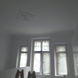 VPBud - Białe wnętrze w trakcie prac: świeżo pomalowane ściany, zabezpieczone folią okna, drewniana drabina i dekoracyjny element na suficie.