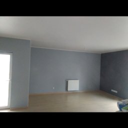 VPBud - Pomalowane na szaro wnętrze pokoju z drewnianą podłogą, białym grzejnikiem i drzwiami balkonowymi po lewej stronie.