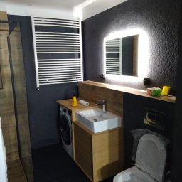 VPBud - Nowoczesna łazienka z czarną teksturowaną ścianą, podświetlanym lustrem, umywalką na drewnianej szafce obok pralki oraz białym grzejnikiem drabinkowym.