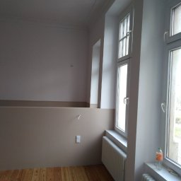 VPBud - Pomieszczenie po malowaniu ścian na dwa kolory: biały u góry i beżowy u dołu, z drewnianą podłogą, białymi grzejnikami i oknami.