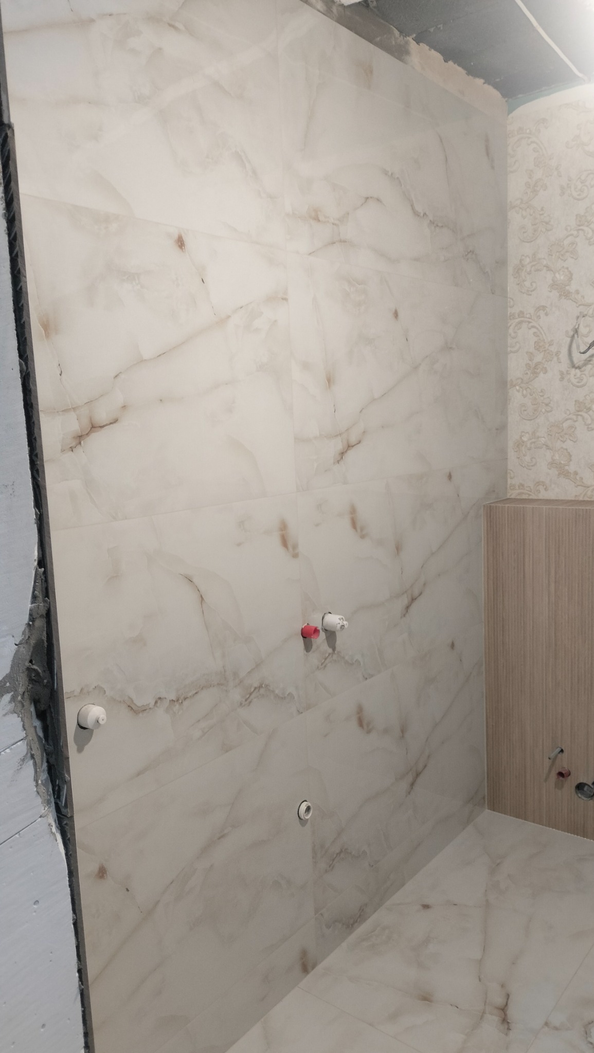Ściana i podłoga w trakcie wykańczania, wyłożone płytkami imitującymi marmur w odcieniach bieli i beżu, widoczne przyłącza wodne z czerwonym i białym elementem, fragment ściany z tapetą w kwiatowy...