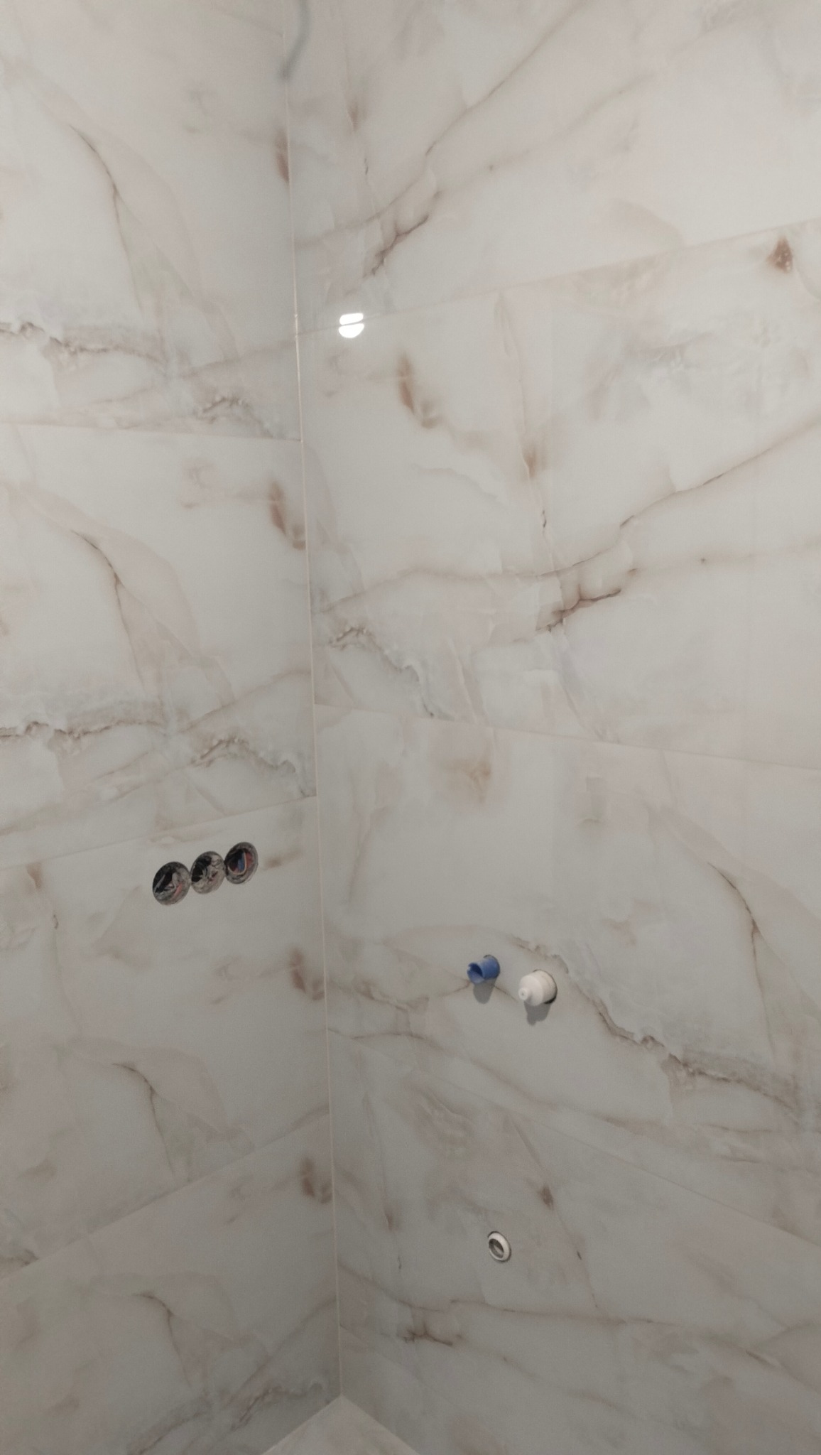 Ściana w łazience wykończona płytkami imitującymi marmur, widoczne otwory na baterię, przyłącza wodne i odpływ w podłodze, etap prac wykończeniowych.