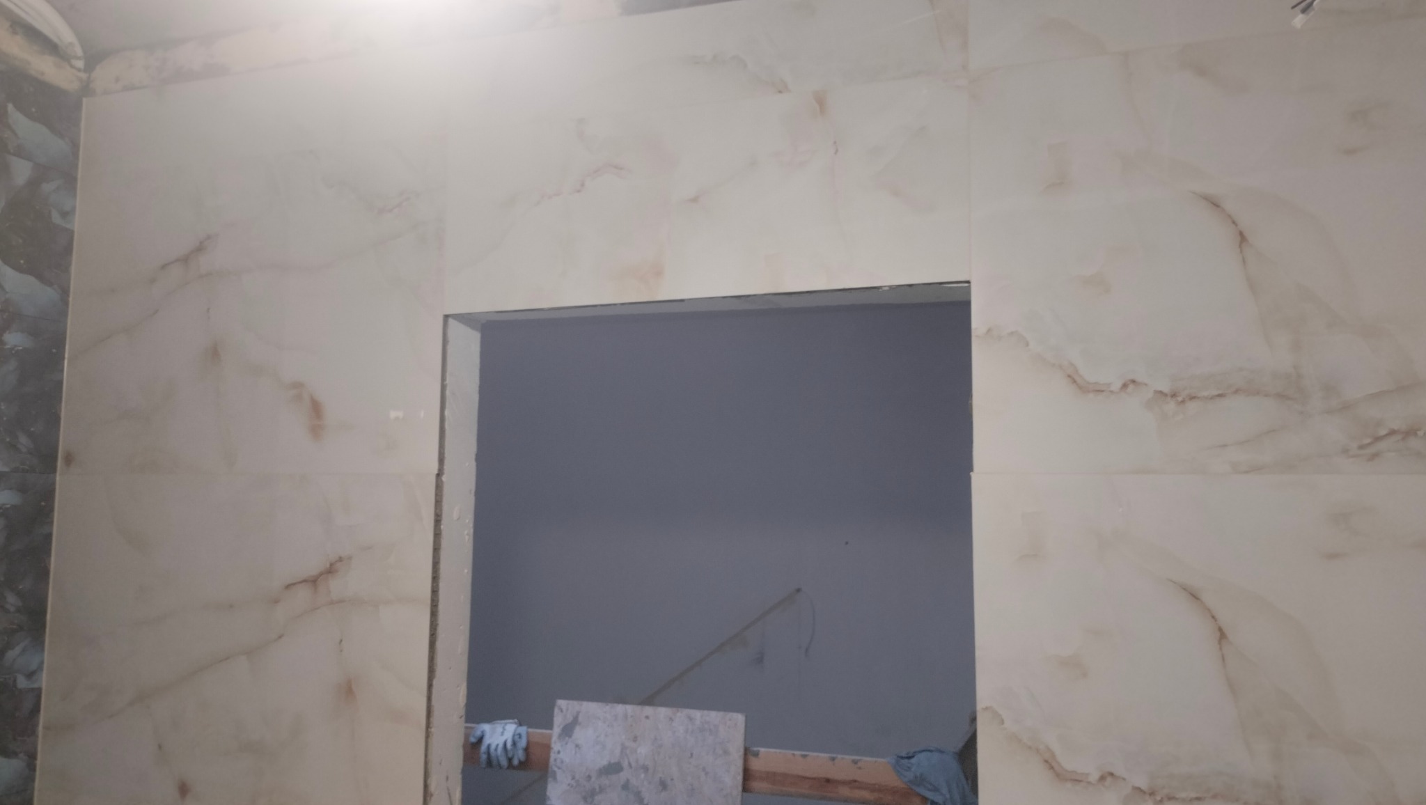Ściana wyłożona jasnymi płytkami imitującymi marmur z widocznym otworem drzwiowym w trakcie prac wykończeniowych, z fragmentem drewnianej konstrukcji i rękawicami roboczymi w tle.