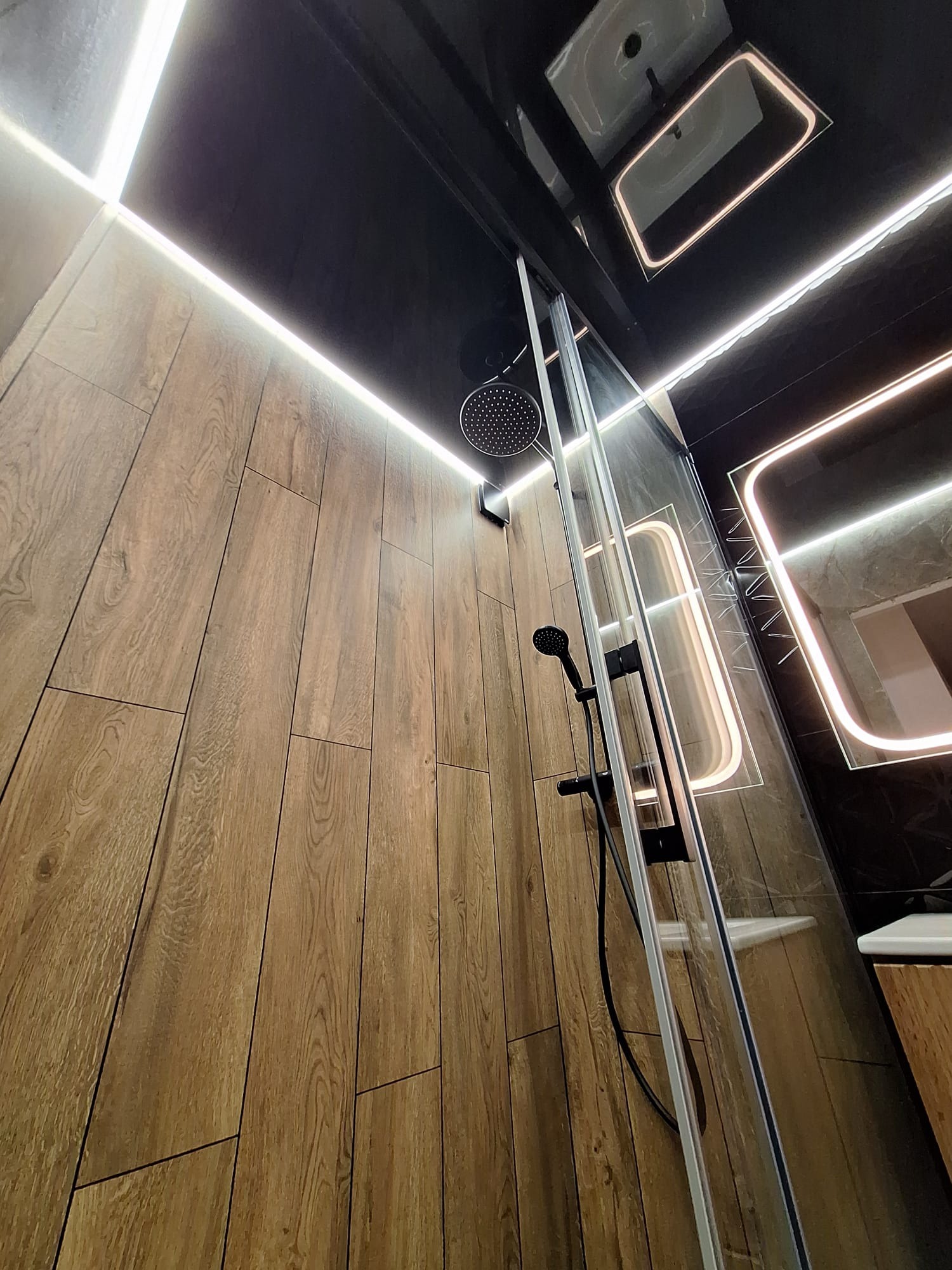 Nowoczesna kabina prysznicowa z deszczownicą, drewnopodobne płytki na ścianie, czarny sufit z oświetleniem LED, lustro podświetlone od tyłu.