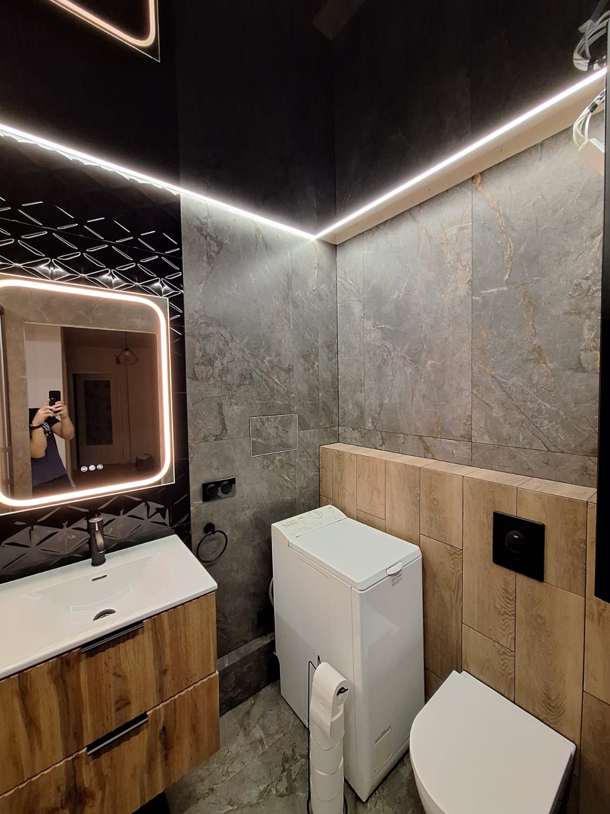 Nowoczesna łazienka z umywalką na szafce z drewnopodobnymi frontami, podświetlanym lustrem, szarą marmurkową glazurą, czarnym sufitem z oświetleniem LED, pralką i toaletą.