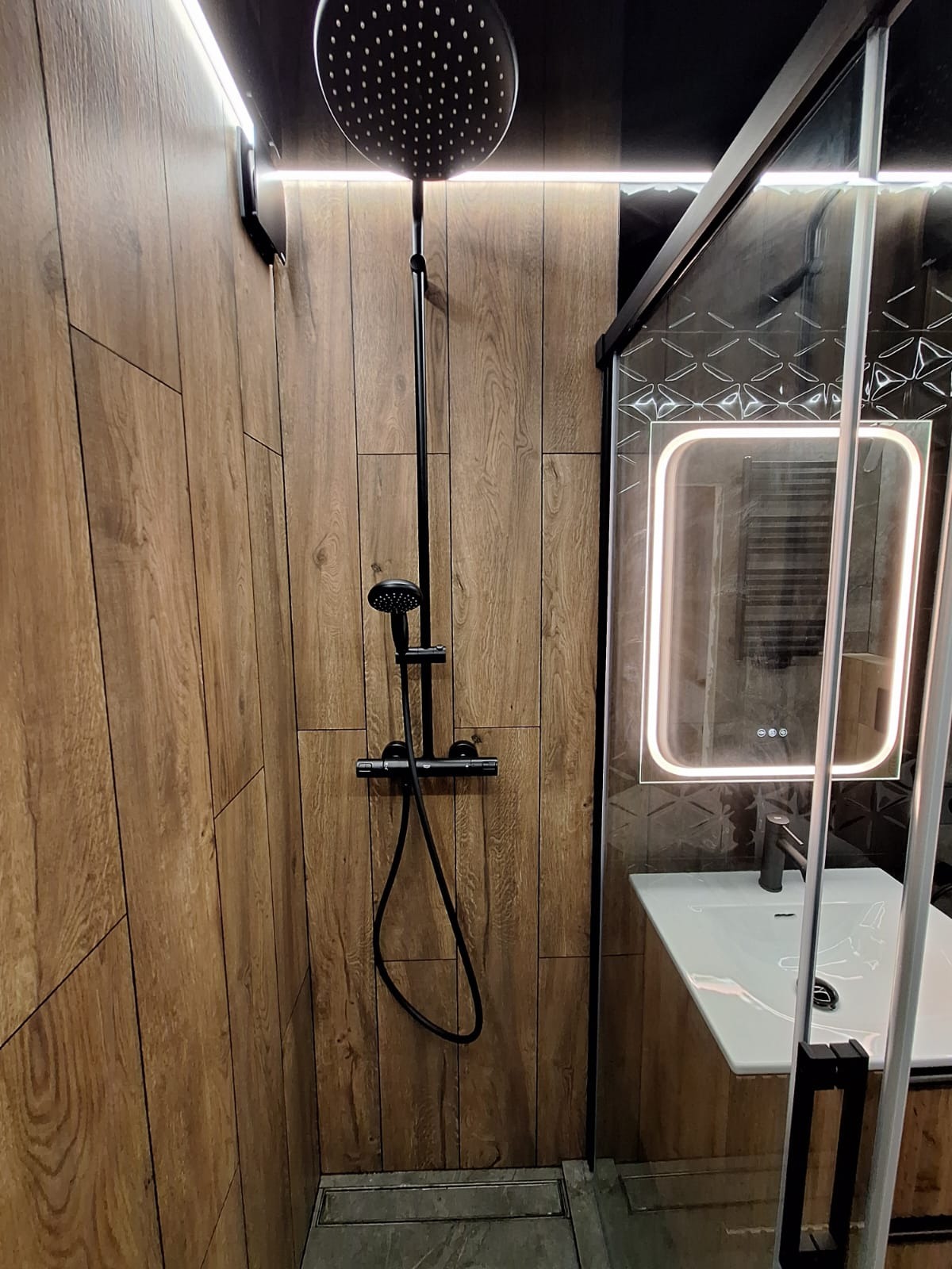 Nowoczesna kabina prysznicowa z deszczownicą i panelem ściennym imitującym drewno, oświetlone lustro z umywalką w tle.