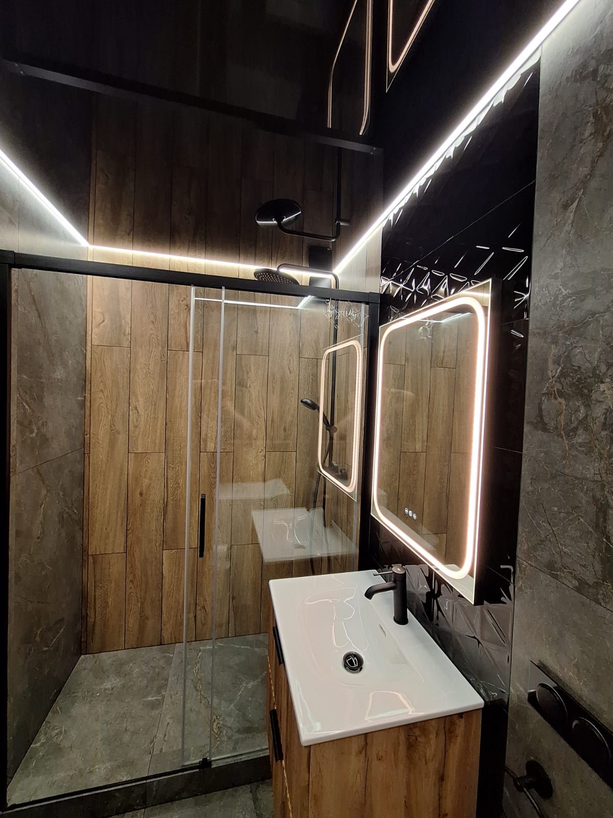 Nowoczesna łazienka z kabiną prysznicową typu walk-in, umywalką nablatową na szafce z drewnopodobnym dekorem, podświetlanymi lustrami i czarną armaturą.