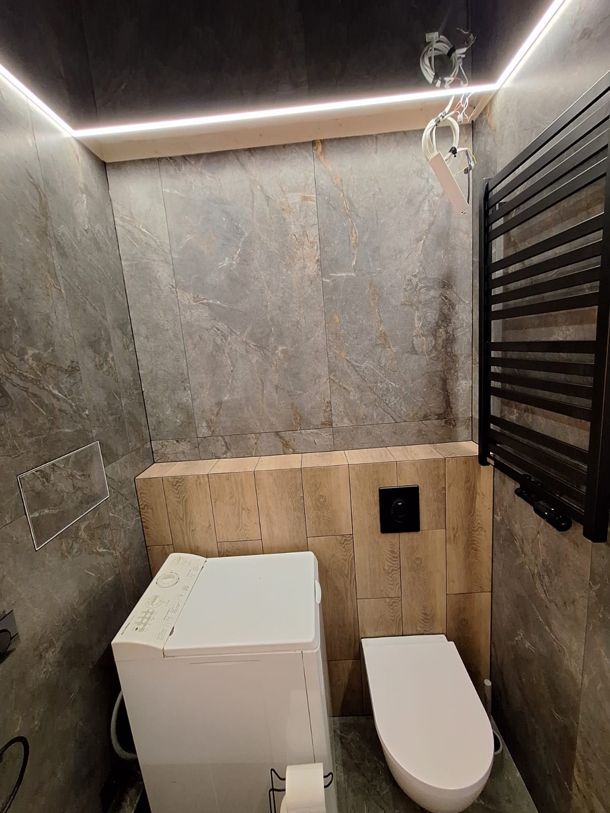 Ukończona łazienka z płytkami imitującymi marmur, podświetleniem LED, grzejnikiem drabinkowym, pralką i podwieszaną toaletą; widoczne niedokończone podłączenie oświetlenia.