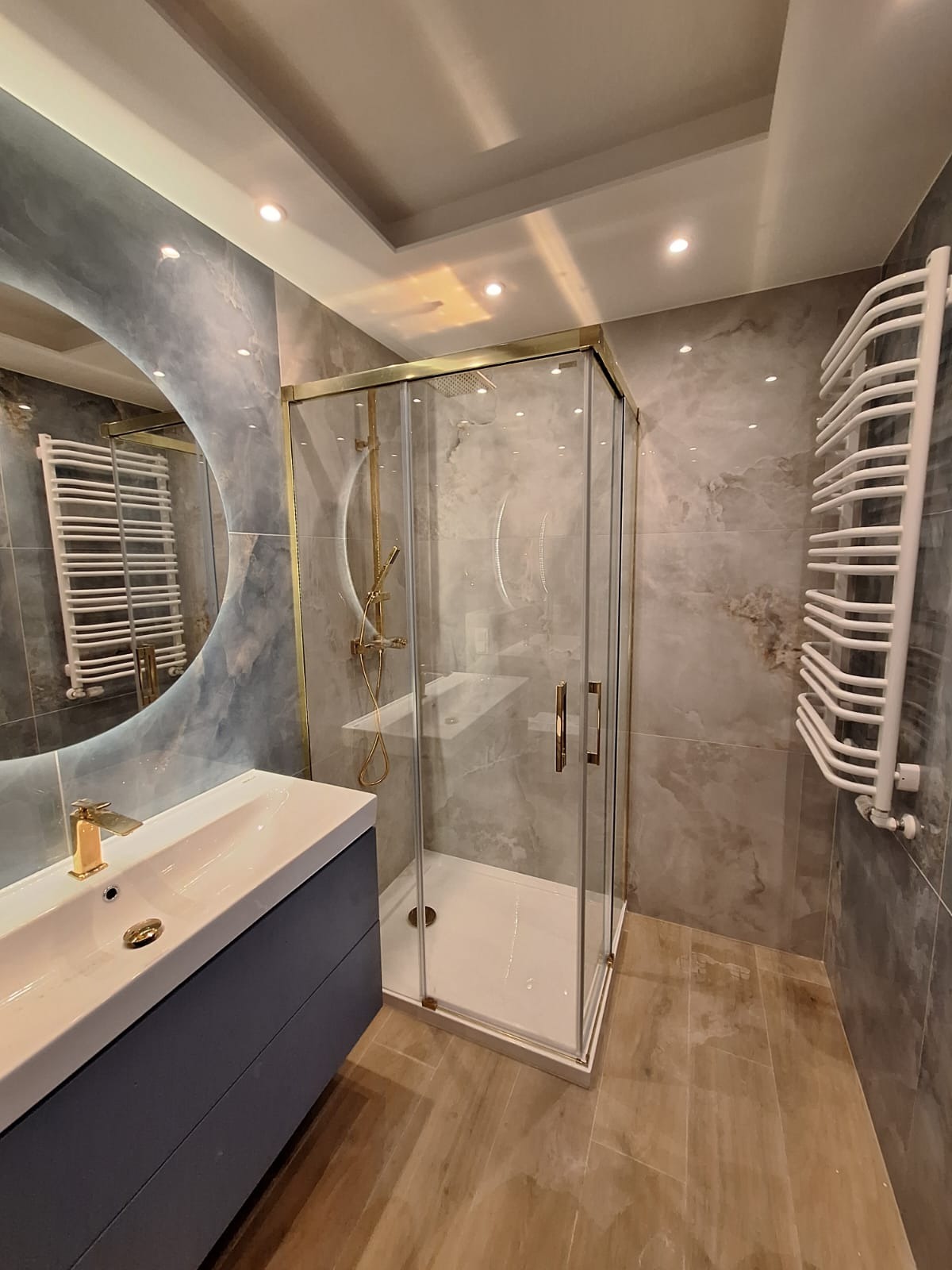 Nowoczesna łazienka z kabiną prysznicową w złotej ramie, umywalką na szafce w odcieniach szarości i okrągłym lustrem z podświetleniem, grzejnik drabinkowy na ścianie z szarych płytek imitujących...