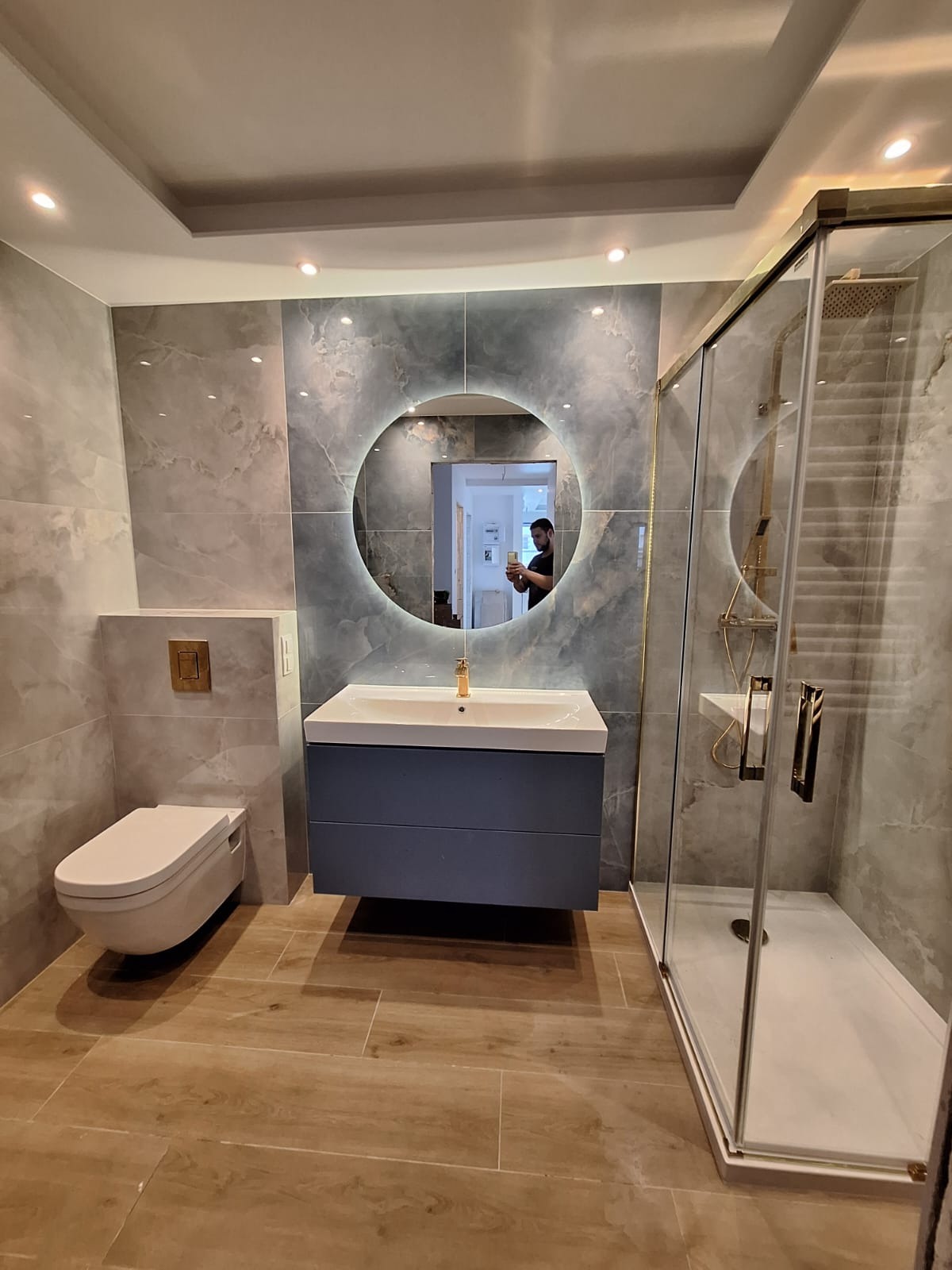 Nowoczesna łazienka we Wrocławiu z szarymi płytkami imitującymi marmur, podwieszana toaleta, umywalka z szafką w kolorze granatowym i okrągłe lustro z podświetleniem LED oraz kabina prysznicowa ze...