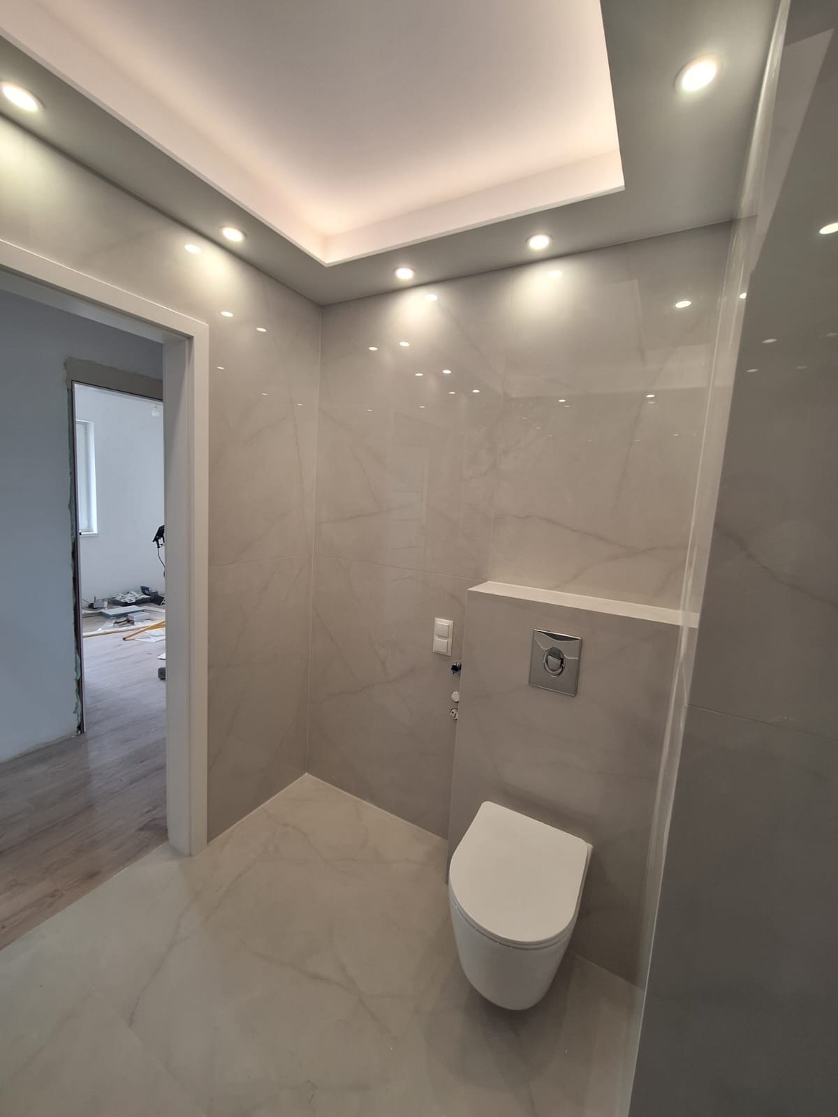 Nowoczesna łazienka z beżowymi płytkami imitującymi marmur, podwieszana toaleta, oświetlenie punktowe w suficie podwieszanym i widok na remontowane pomieszczenie obok.