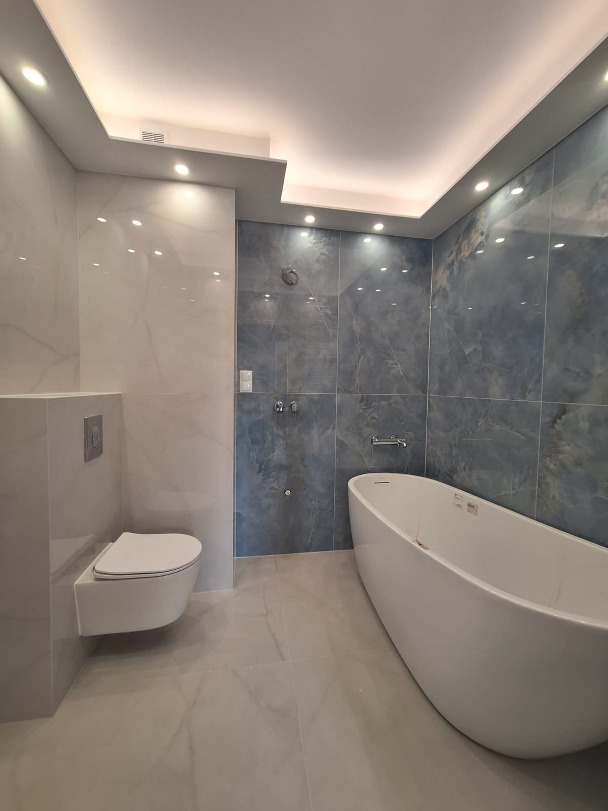 Nowoczesna łazienka z podwieszaną toaletą, wolnostojącą wanną i marmurowymi płytkami w odcieniach szarości i błękitu, oświetlona punktowymi lampami w suficie.
