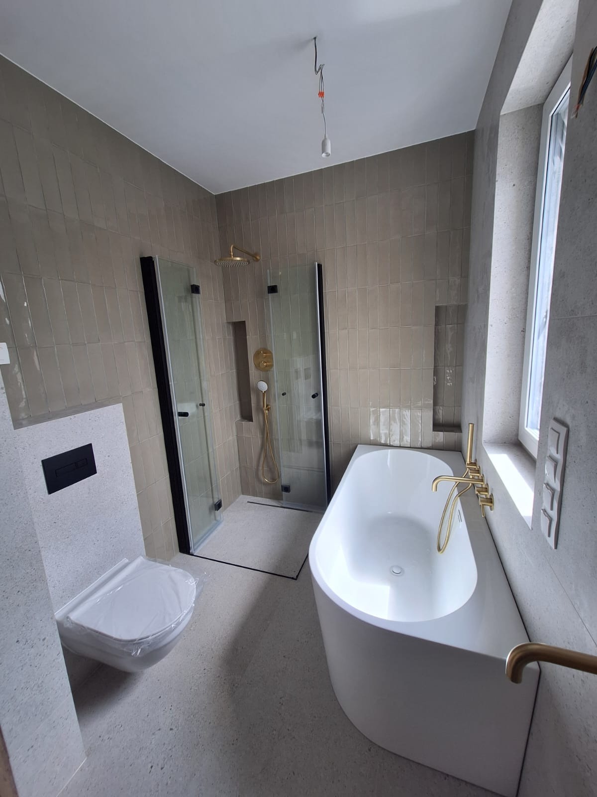 Nowoczesna łazienka z beżowymi płytkami, kabiną prysznicową z czarnymi ramami, białą wanną wolnostojącą z złotą armaturą i wiszącą toaletą z czarnym przyciskiem, całość utrzymana w minimalistycznym...