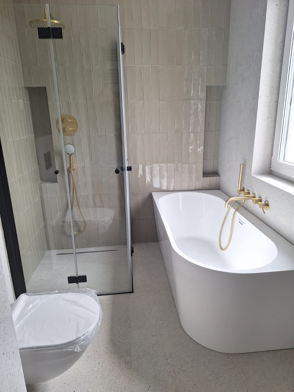 Nowoczesna łazienka z beżowymi, pionowo ułożonymi płytkami, kabiną prysznicową typu walk-in z czarną ramą, złotą armaturą i białą wanną.