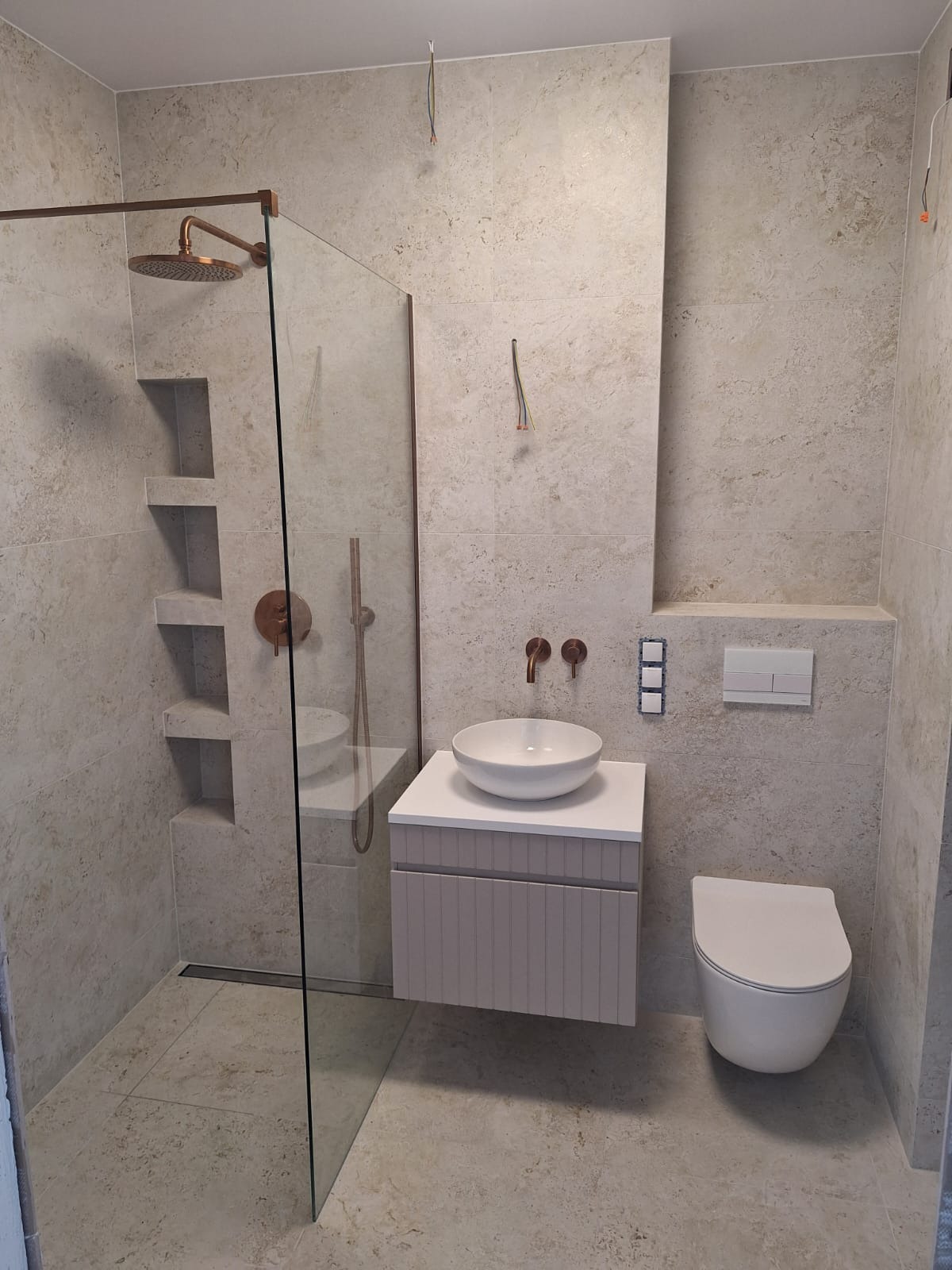 Nowoczesna łazienka z beżowymi płytkami imitującymi kamień, kabiną prysznicową typu walk-in z miedzianymi elementami, umywalką nablatową i podwieszaną toaletą.