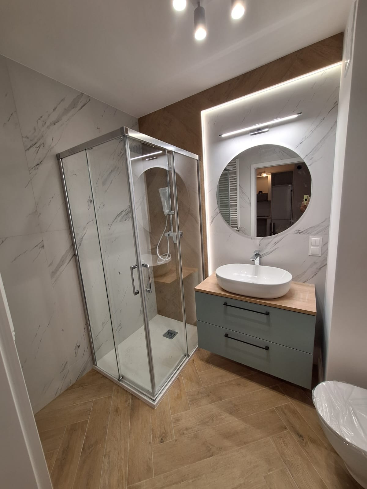 Nowoczesna łazienka z kabiną prysznicową, okrągłym lustrem z podświetleniem LED, umywalką nablatową na szafce z szufladami w kolorze pastelowej zieleni i podłogą z płytek imitujących drewno...