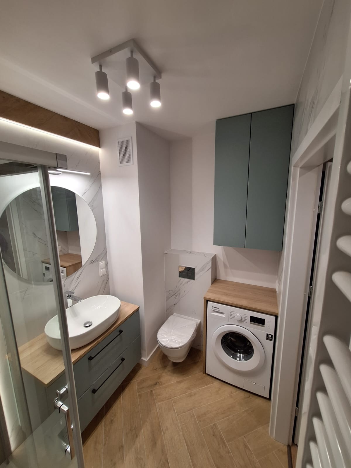 Nowoczesna łazienka z drewnopodobną podłogą, umywalką nablatową na szafce z szufladami, toaletą w zabudowie, pralką pod blatem i szafką wiszącą w odcieniach szarości i bieli.