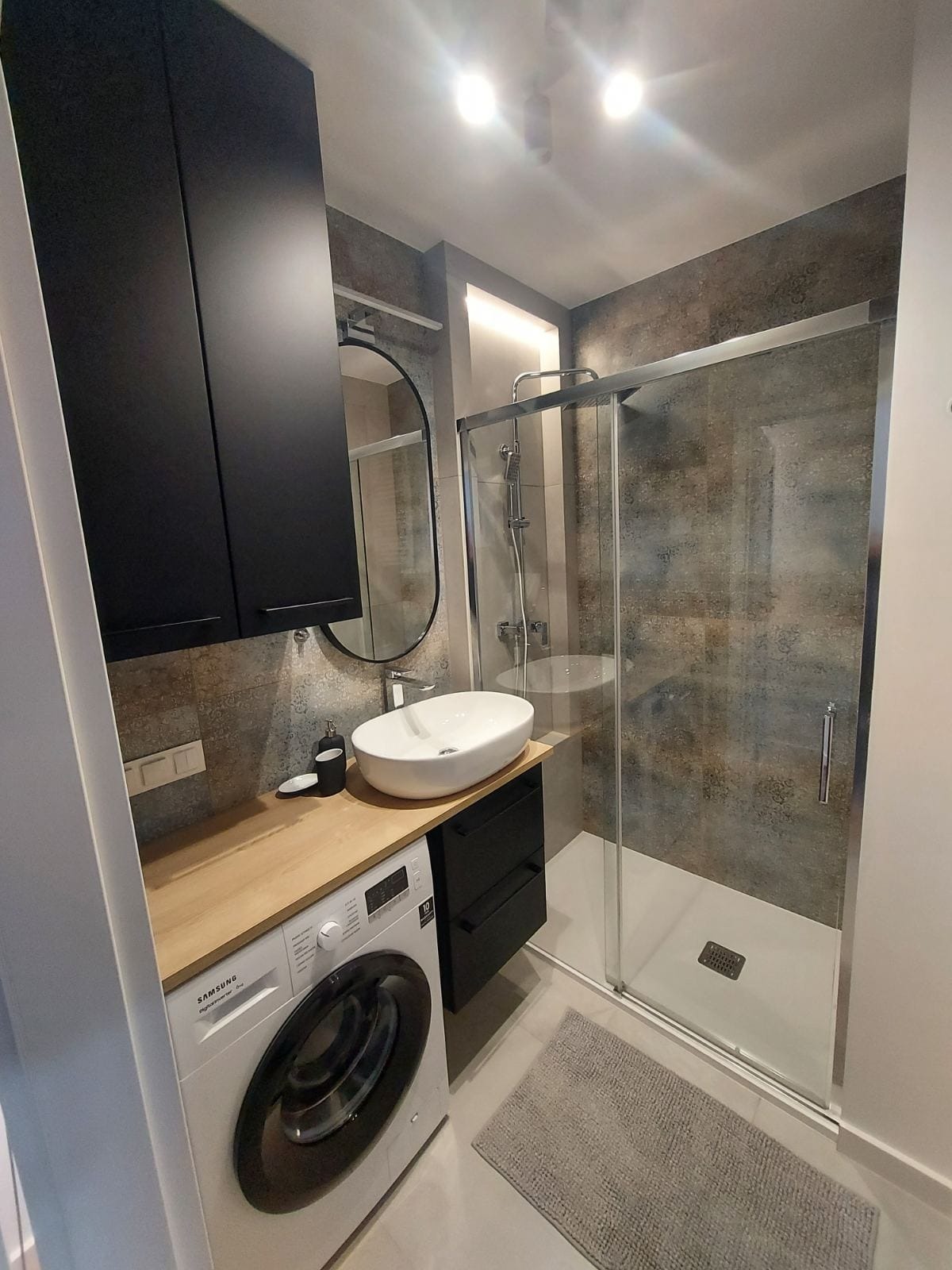 Nowoczesna łazienka z kabiną prysznicową typu walk-in, umywalką nablatową owalną, pralką Samsung pod blatem i czarnymi szafkami wiszącymi, oświetlona punktowymi lampami sufitowymi.