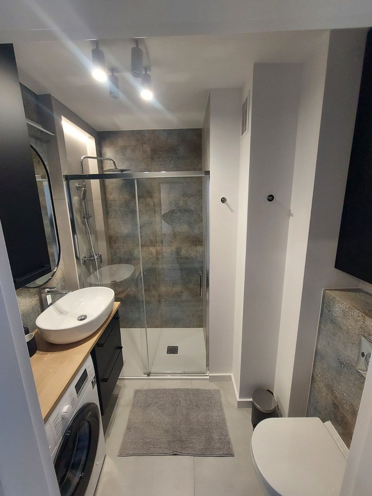 Nowoczesna łazienka z kabiną prysznicową, umywalką nablatową na szafce z pralką, szara wycieraczka i toaleta.