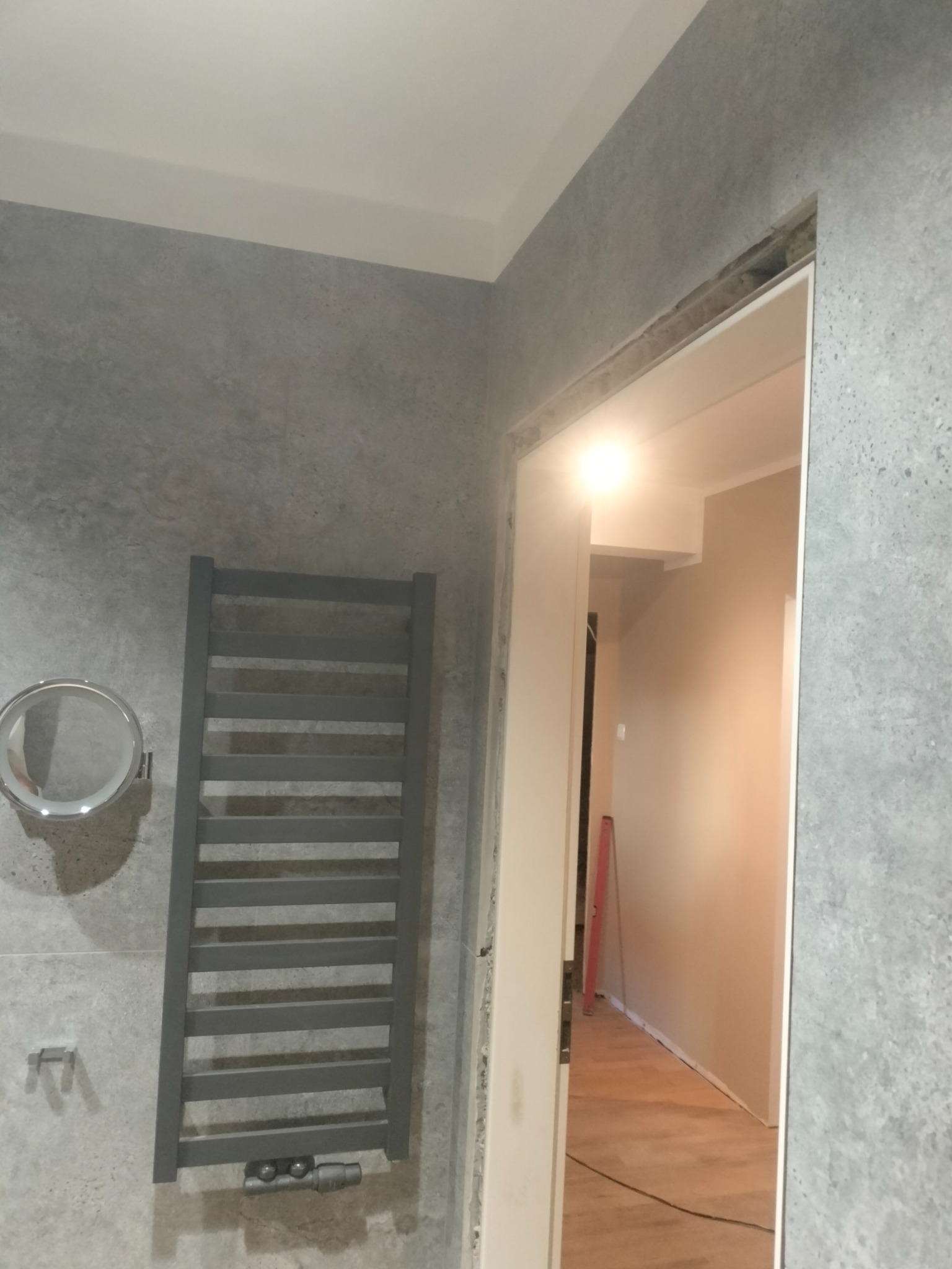 Szara łazienka z grzejnikiem drabinkowym i okrągłym lustrem, widok na korytarz z drewnianą podłogą przez otwarte drzwi bez ościeżnicy.