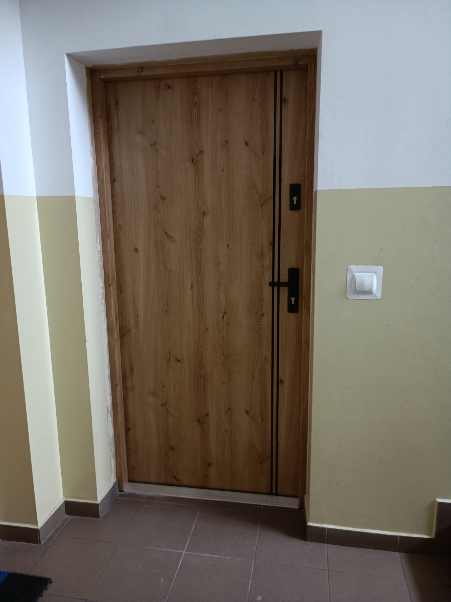 Zamknięte drzwi wejściowe z drewnopodobną okleiną i czarnymi, pionowymi paskami, zamontowane w bloku mieszkalnym, widoczny włącznik światła na ścianie.