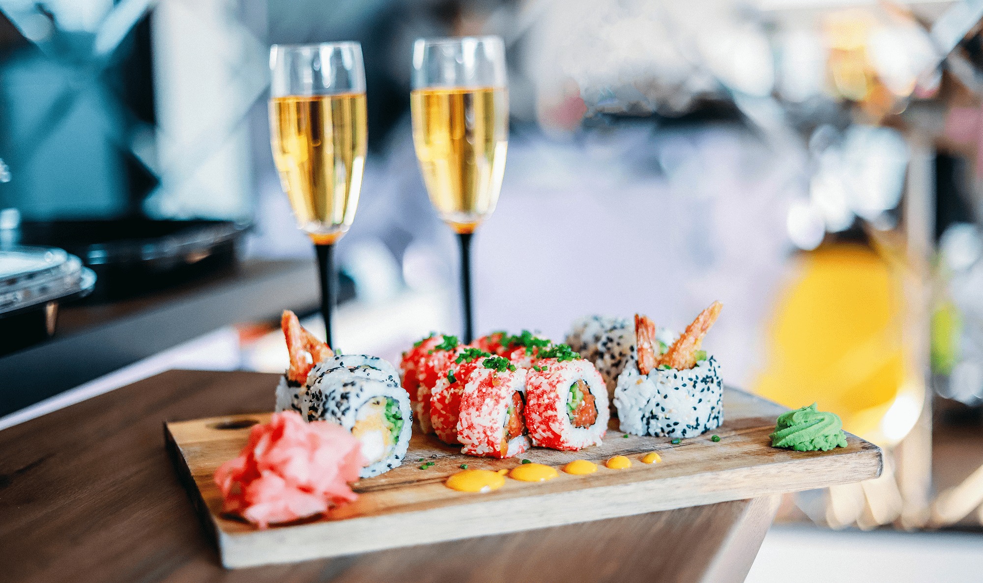 Elegancko podane sushi z krewetkami i wasabi na drewnianej desce, w tle dwa kieliszki z szampanem i delikatny bokeh.