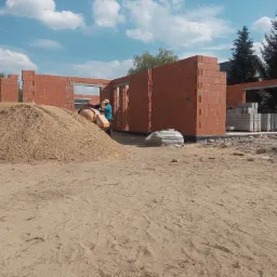 Budowa domu z czerwonej cegły ceramicznej Porotherm: widoczny etap murowania ścian, stos piasku, betoniarka i pracownik w tle.