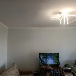 Jasne wnętrze pokoju z błękitnymi ścianami, nowoczesną lampą sufitową w kształcie abstrakcyjnych linii, telewizorem z wyświetlanym obrazem drzew i częściowo widoczną kanapą w paski.