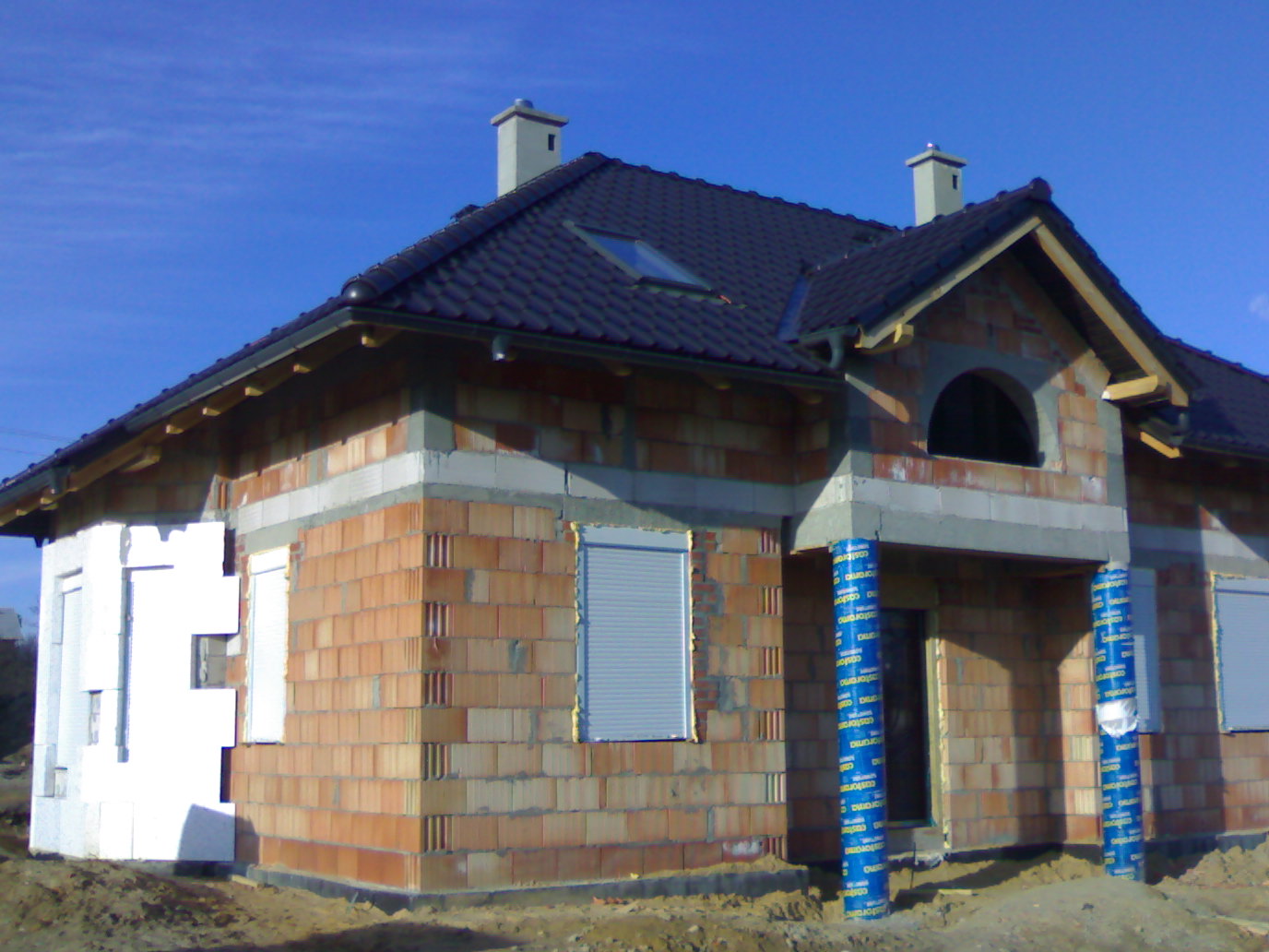 Budowa domu jednorodzinnego w stanie surowym otwartym; widoczne ściany z cegły, częściowo ocieplone styropianem, zamontowane rolety zewnętrzne, dach z czarnej dachówki i dwa kominy.