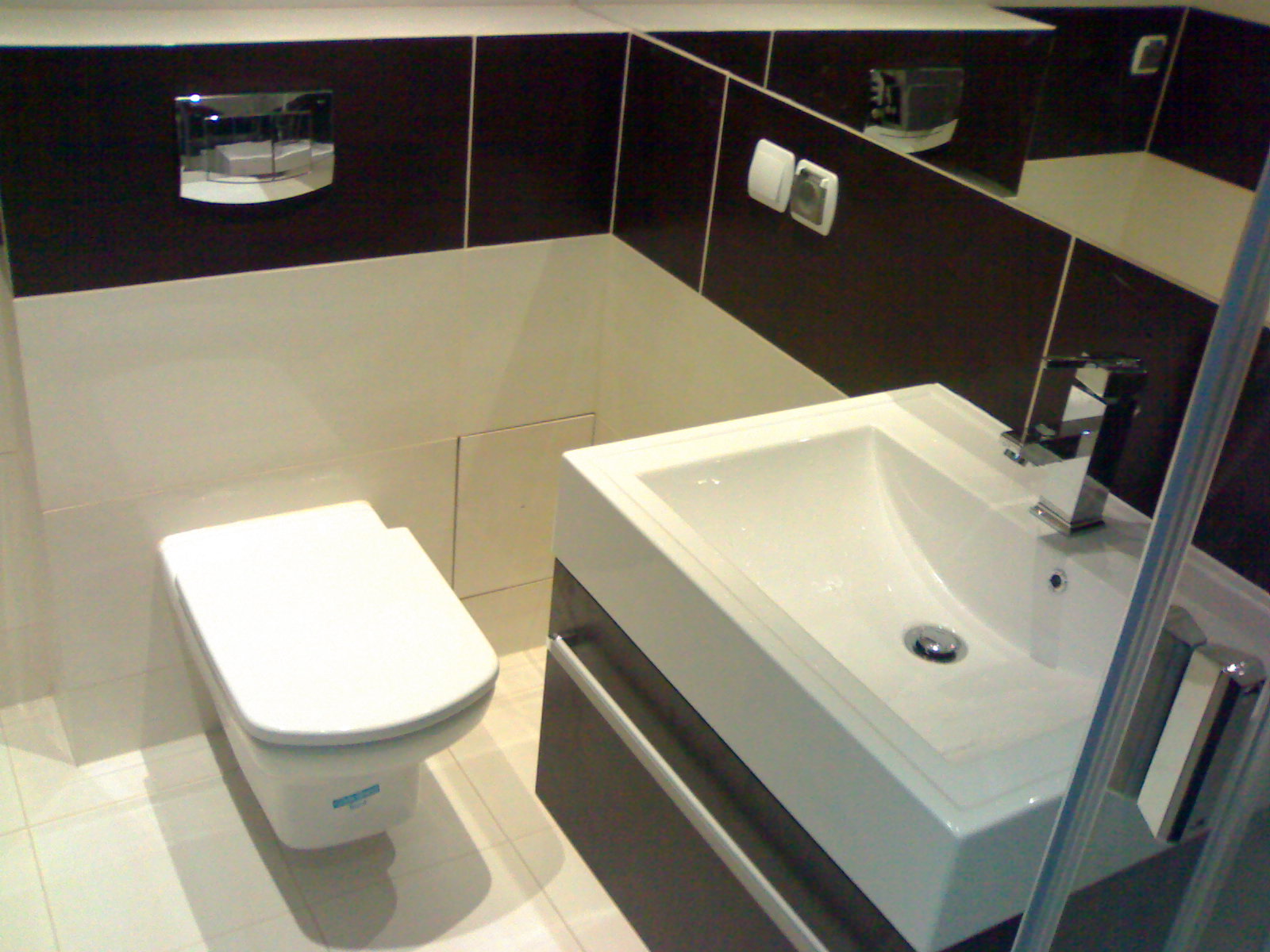 Nowoczesna łazienka z białymi i brązowymi płytkami, wisząca toaleta, umywalka z szafką i chromowaną baterią.