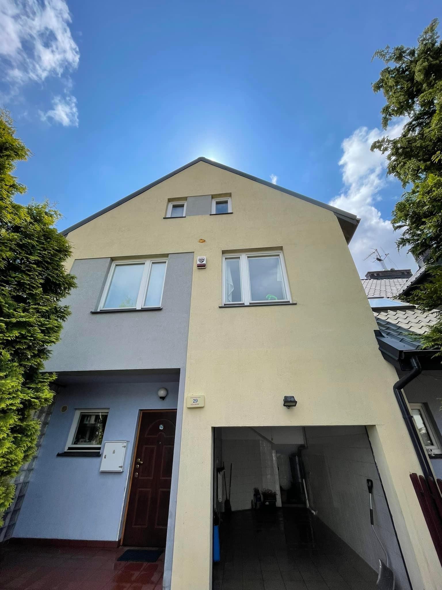 Dwukondygnacyjny dom z elewacją w odcieniach beżu i szarości, widziany z dołu pod kątem, z widocznymi oknami, drzwiami wejściowymi i garażem, na tle błękitnego nieba z chmurami.