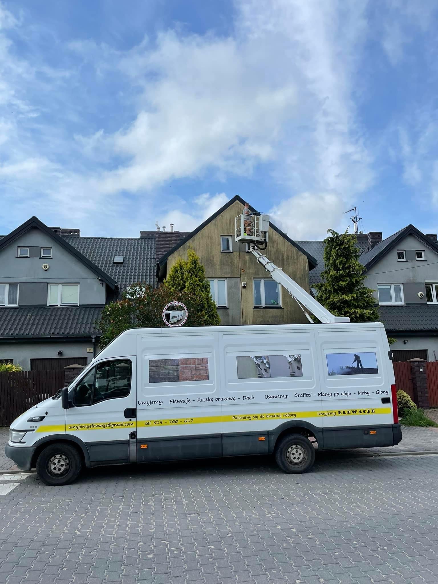 Pracownik na podnośniku koszowym w Nadmie czyści elewację budynku. Na pierwszym planie biały samochód dostawczy z reklamą firmy oferującej usługi czyszczenia elewacji, kostki brukowej i dachów.