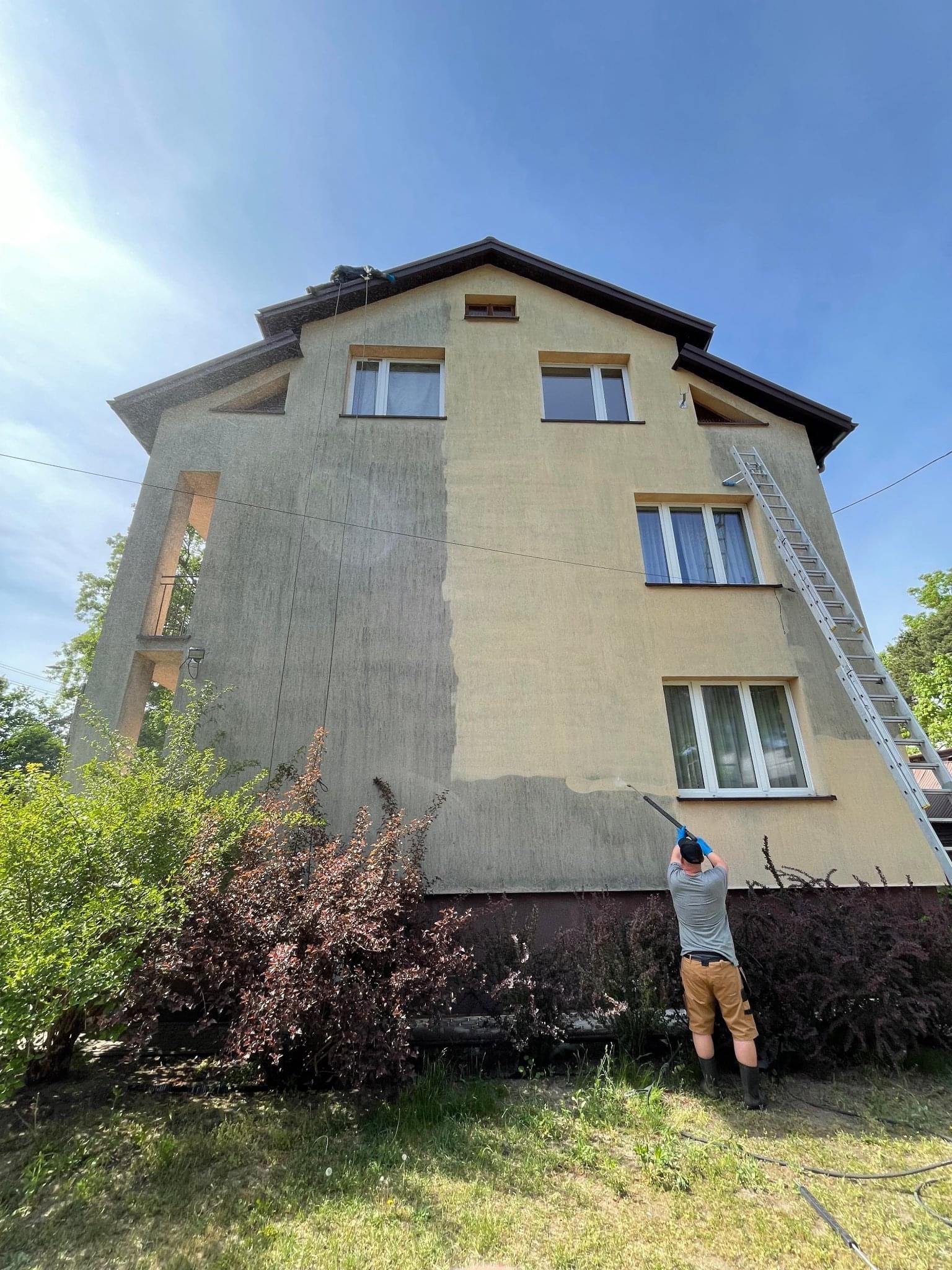 Dwukondygnacyjny dom z częściowo wyczyszczoną elewacją, widoczna różnica w kolorze, pracownik z myjką ciśnieniową na dole i drugi na dachu, drabina oparta o ścianę, krzewy w ogrodzie, błękitne niebo.