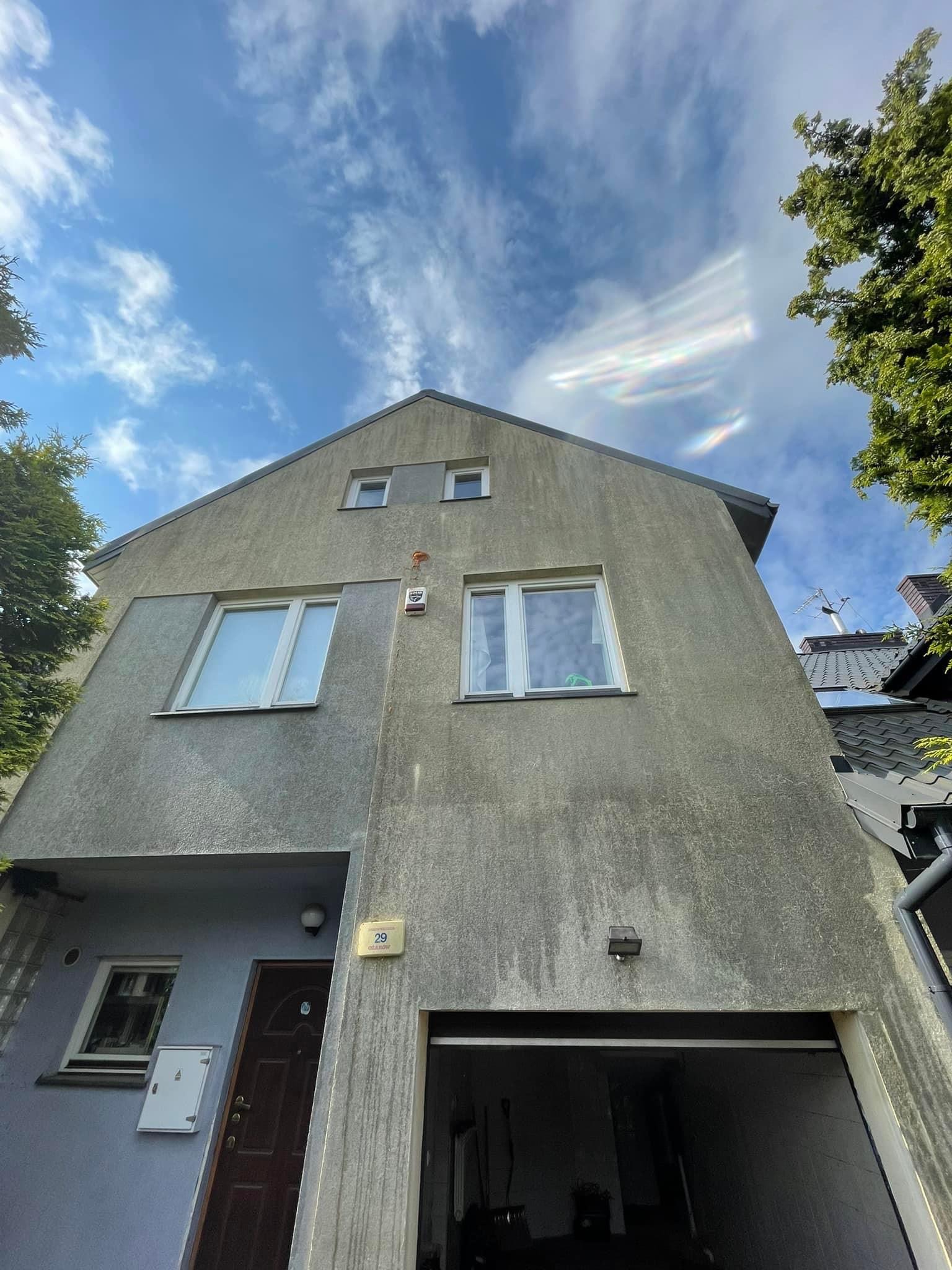 Widok z dołu na szarą elewację dwupiętrowego domu z numerem 29, białymi oknami i otwartym garażem w słoneczny dzień z błękitnym niebem i chmurami.