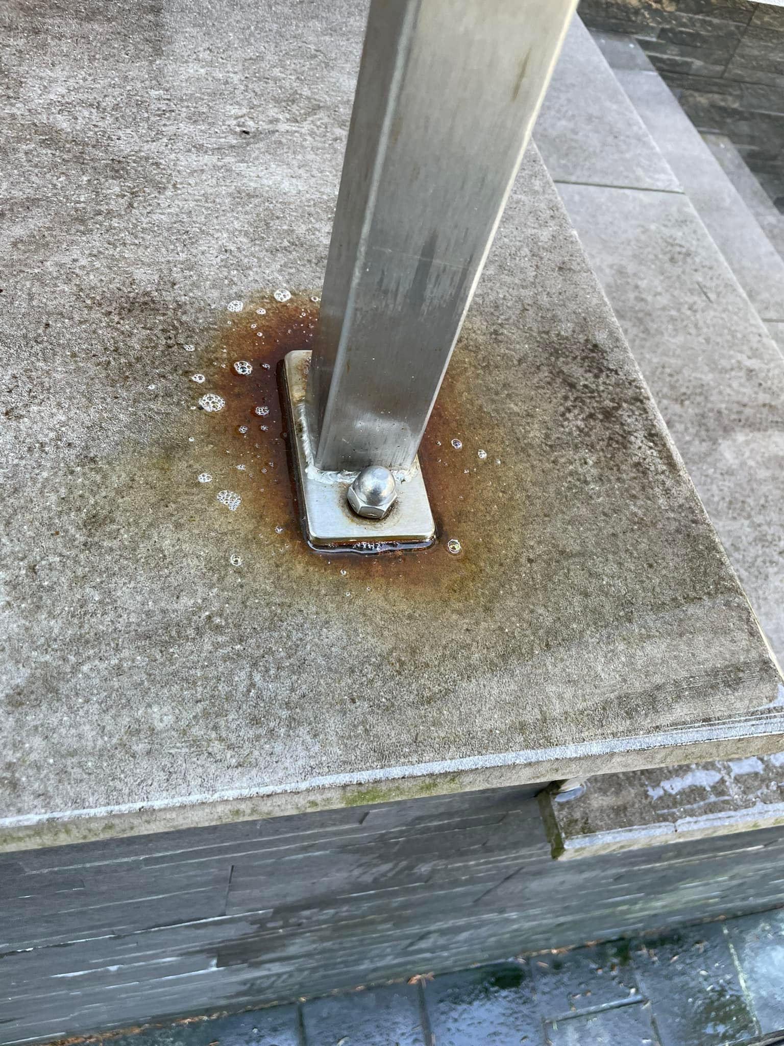 Zacieki rdzy wokół podstawy metalowego słupka mocowanego do kamiennej powierzchni schodów, widoczne ślady czyszczenia.