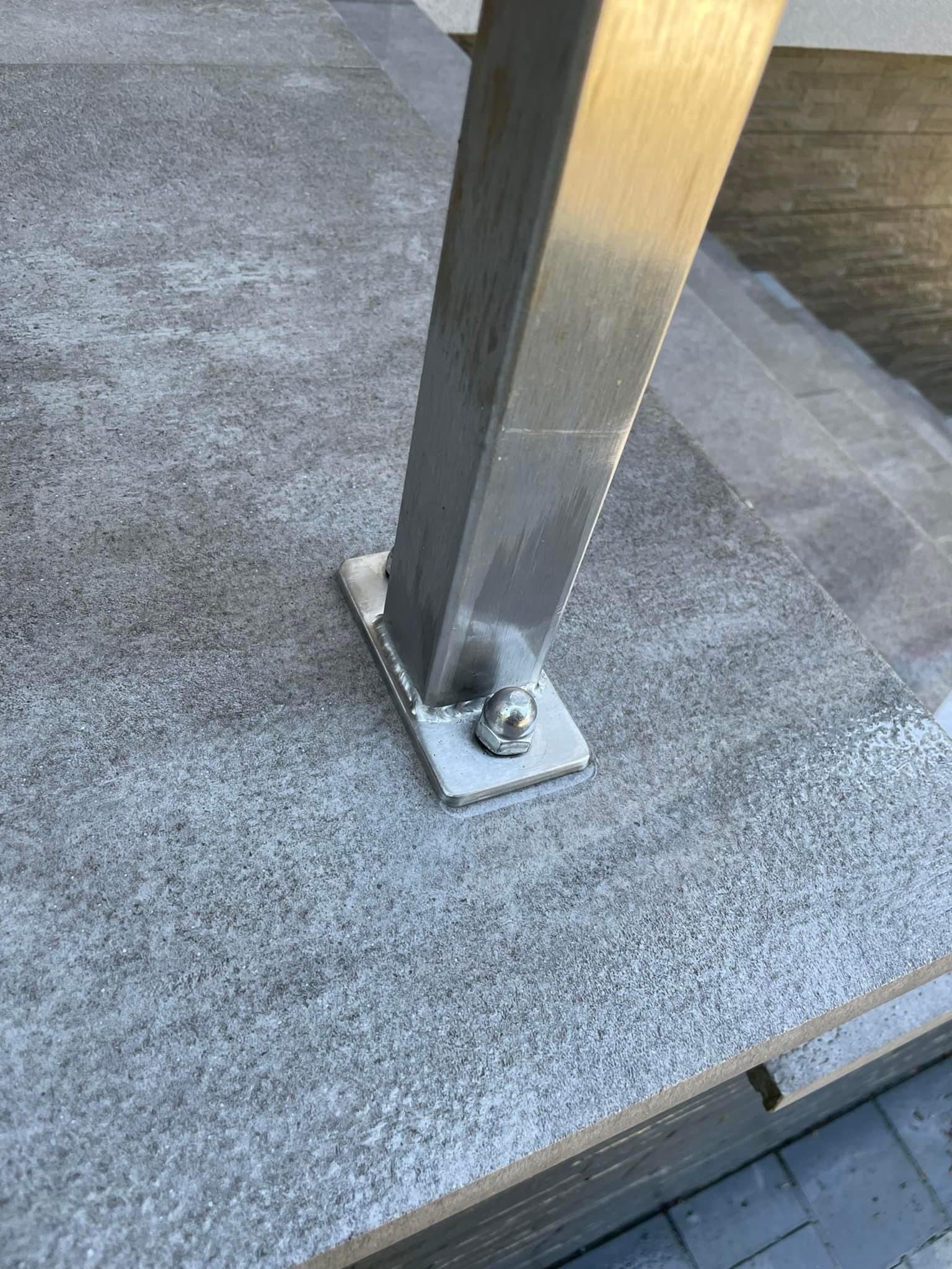 Szczegół mocowania metalowej barierki do kamiennej nawierzchni schodów, widoczne spawy i śruby mocujące.