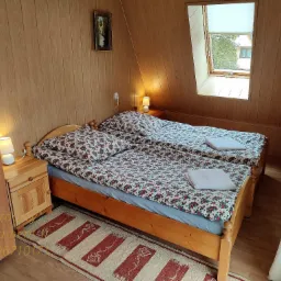 Pokój z dwoma łóżkami pojedynczymi w pensjonacie, drewniane ściany i podłoga, okno dachowe z roletą, lampki nocne na szafkach, dywanik w paski.