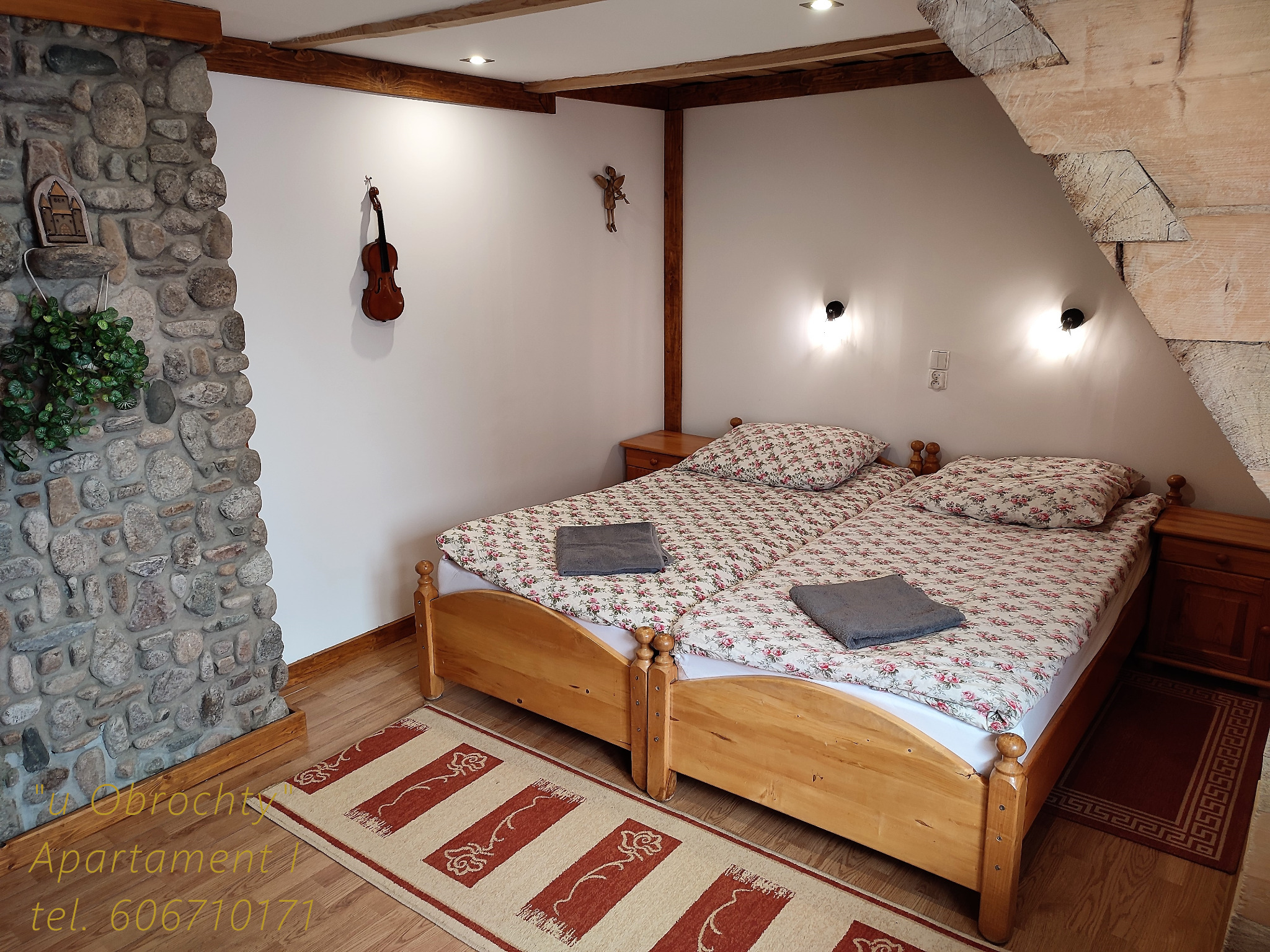 Rustykalna sypialnia z dwoma pojedynczymi łóżkami, kamiennym kominkiem i drewnianymi belkami stropowymi, dekorowana skrzypcami i figurką anioła na ścianie.