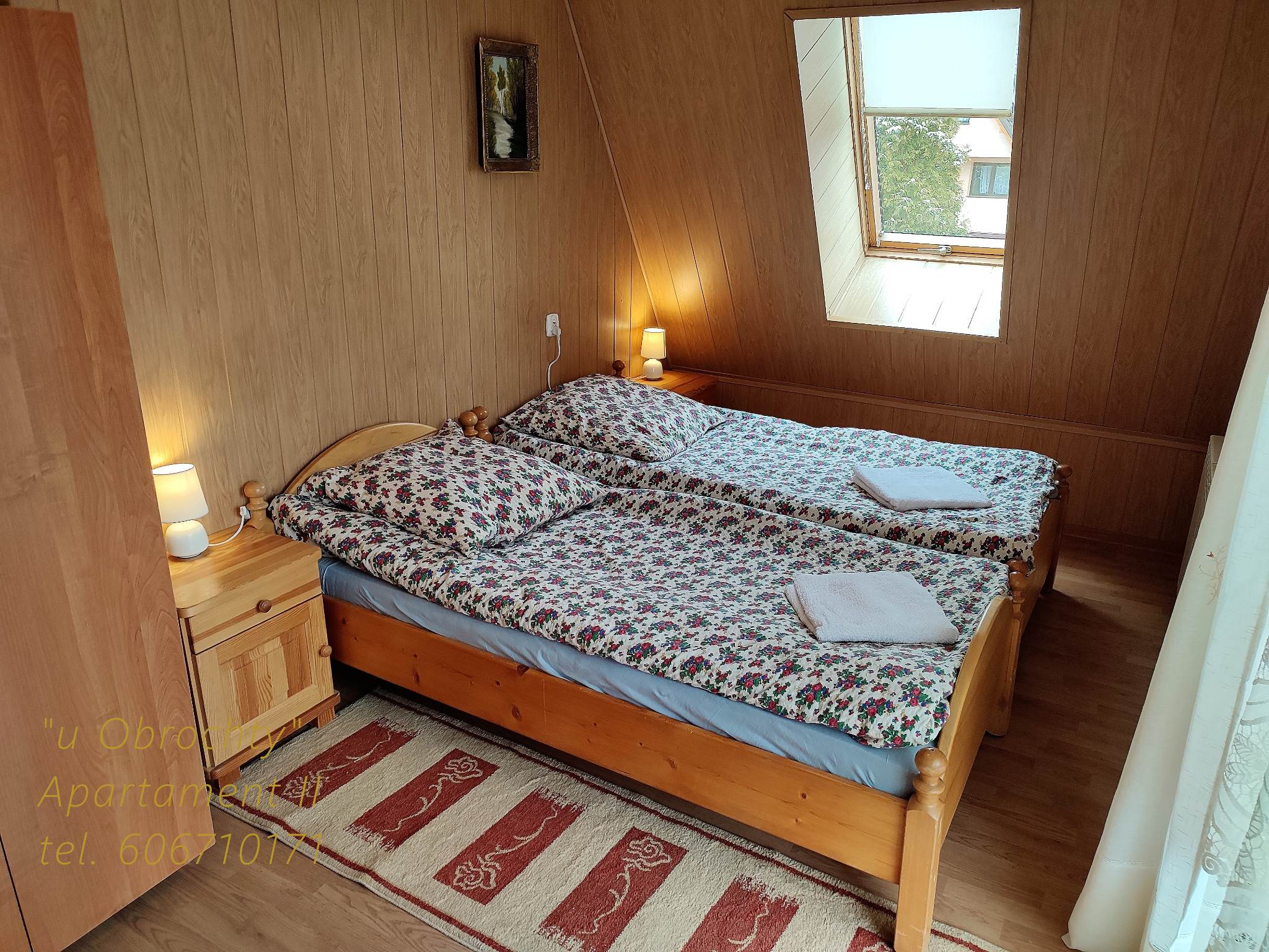 Pokój z dwoma łóżkami pojedynczymi w pensjonacie, drewniane ściany i podłoga, okno dachowe z roletą, lampki nocne na szafkach, dywanik w paski.