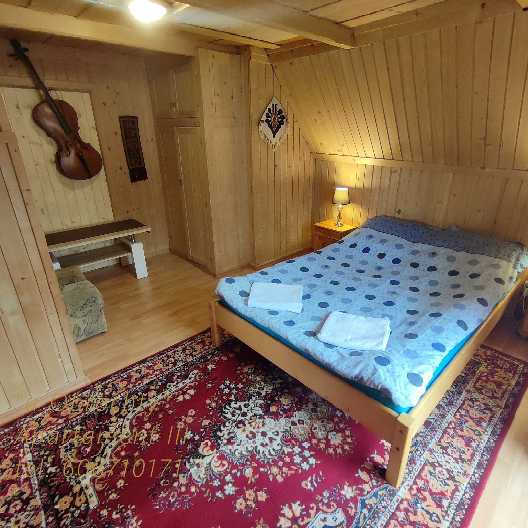 Przytulny pokój w stylu góralskim z drewnianymi ścianami, łóżkiem z niebieską pościelą w kropki, czerwonym dywanem orientalnym, skrzypcami na ścianie i lampką nocną.