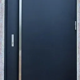 Ciemne skrzydło z długim, pionowym, srebrnym uchwytem zamontowane w surowym otworze drzwiowym z widoczną cegłą i pianką montażową.