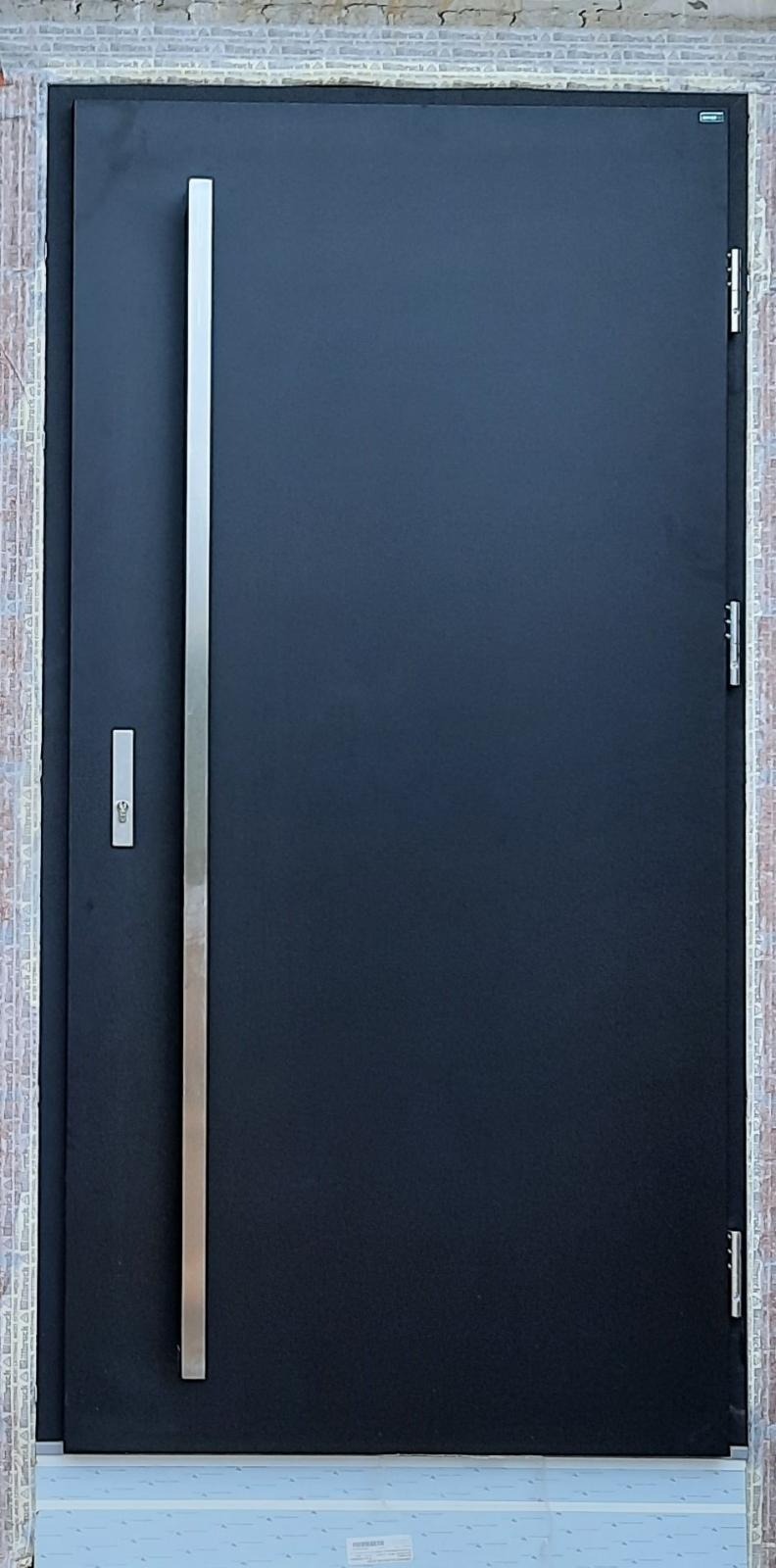 Ciemne skrzydło z długim, pionowym, srebrnym uchwytem zamontowane w surowym otworze drzwiowym z widoczną cegłą i pianką montażową.