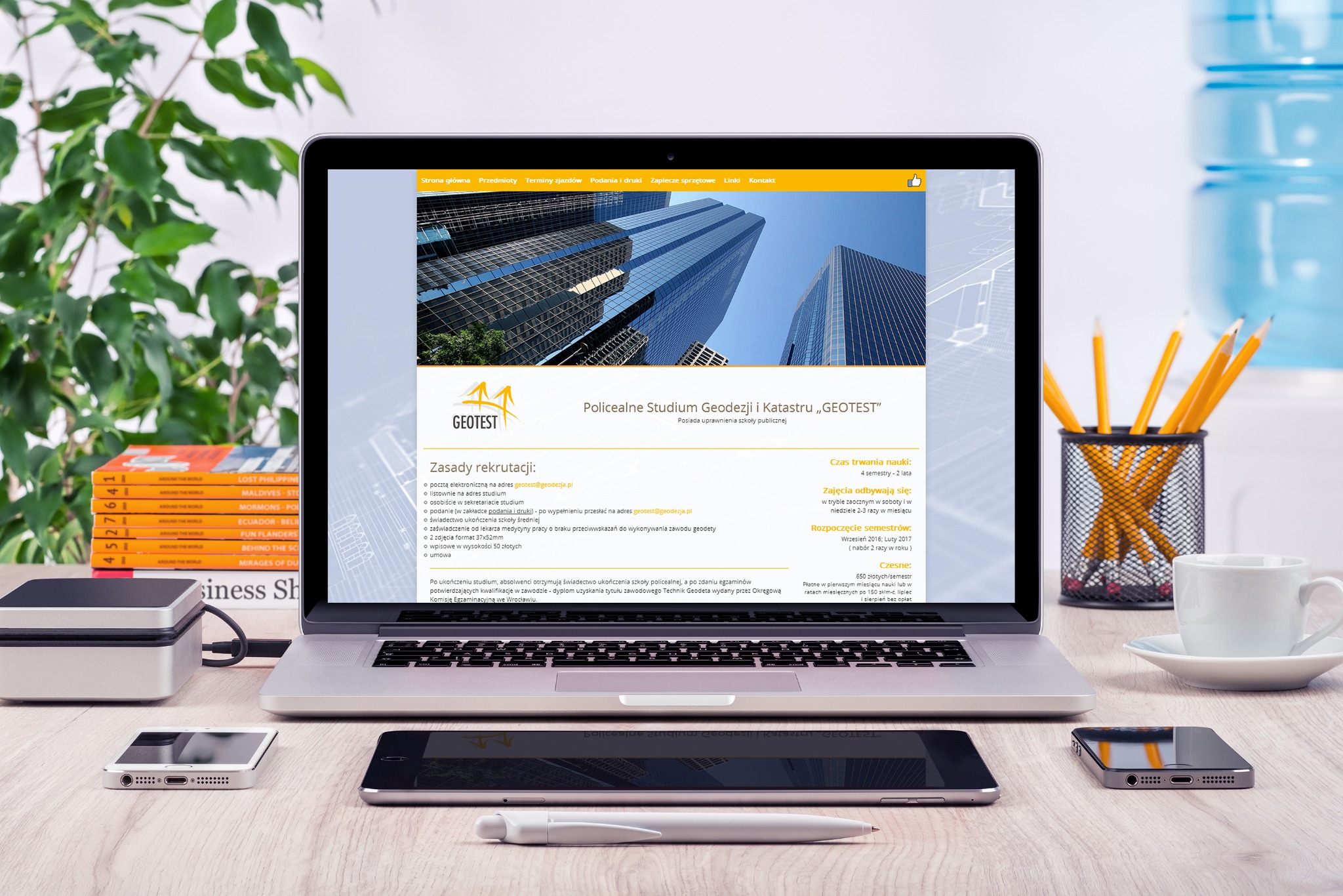 Laptop wyświetlający stronę internetową Policealnego Studium Geodezji i Katastru 'GEOTEST' na biurku z przyborami biurowymi, telefonami, kawą i rośliną doniczkową.
