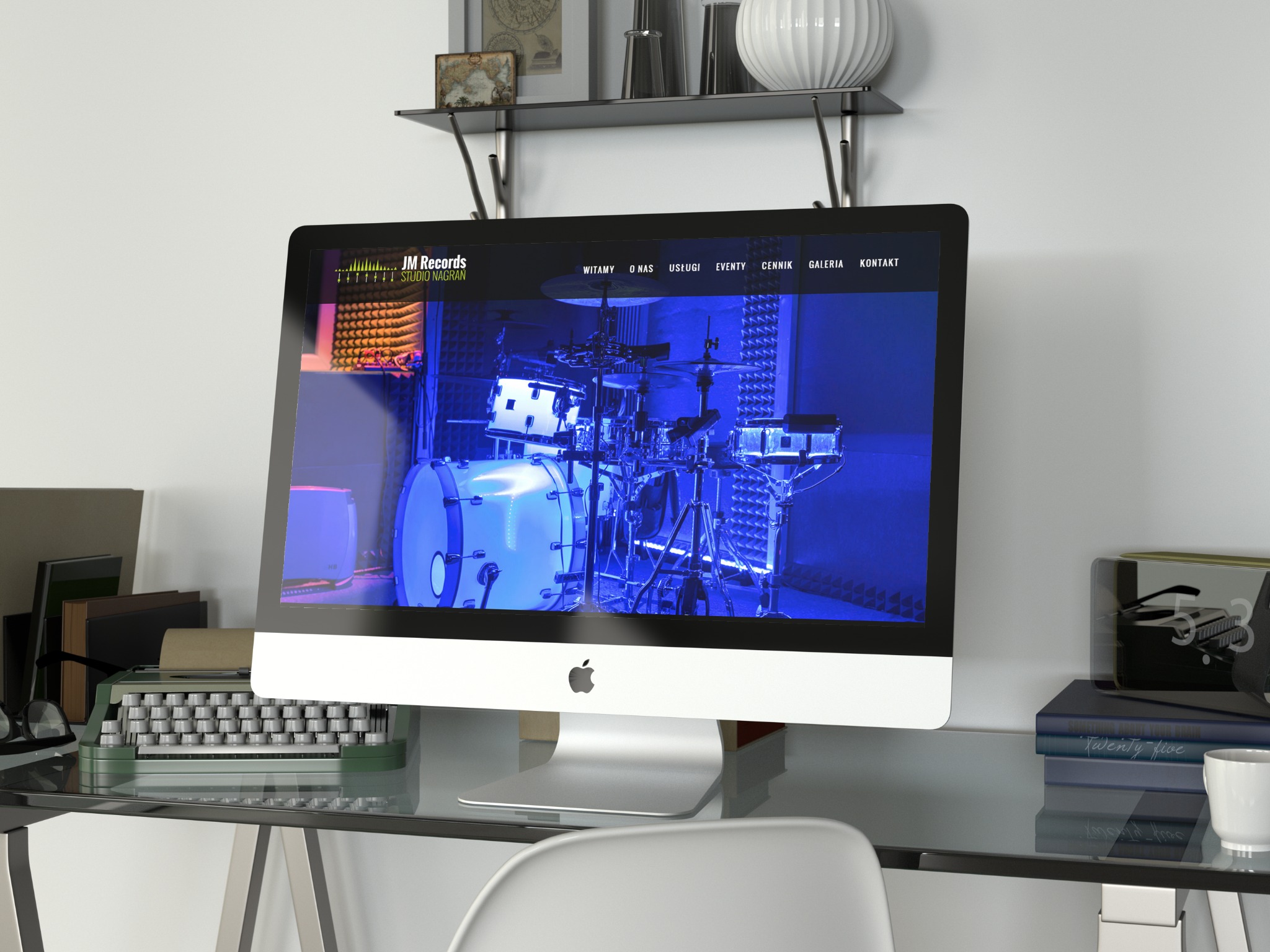 Ekran komputera iMac wyświetlający stronę internetową studia nagrań JM Records z bębnami w tle, na szklanym biurku z maszyną do pisania, książkami i filiżanką, w minimalistycznym wnętrzu.