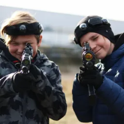Dwoje uśmiechniętych dzieci w kurtkach i czapkach z czujnikami, celujących z pistoletów laserowych w kierunku kamery, na tle jasnej ściany.