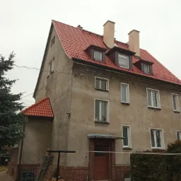 Dwupiętrowy budynek z nowym, czerwonym dachem z dachówki ceramicznej, widoczne okna dachowe i dwa kominy, elewacja w odcieniach szarości, z przodu budynku drzewo iglaste i elementy ogrodzenia.