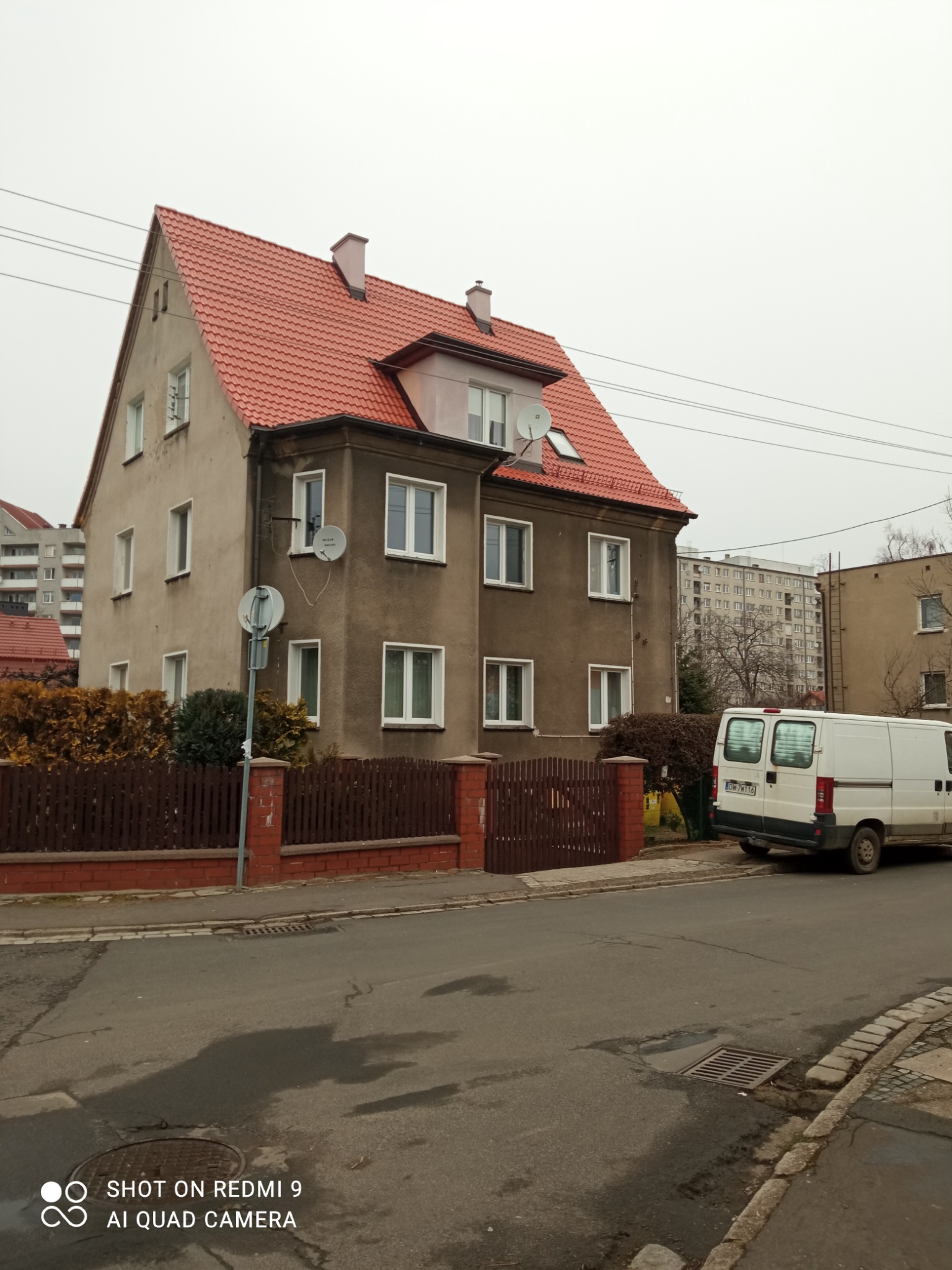 Dwupiętrowy dom z czerwonym dachem z dachówki ceramicznej widziany z ulicy w pochmurny dzień, z zaparkowanym białym vanem w tle.